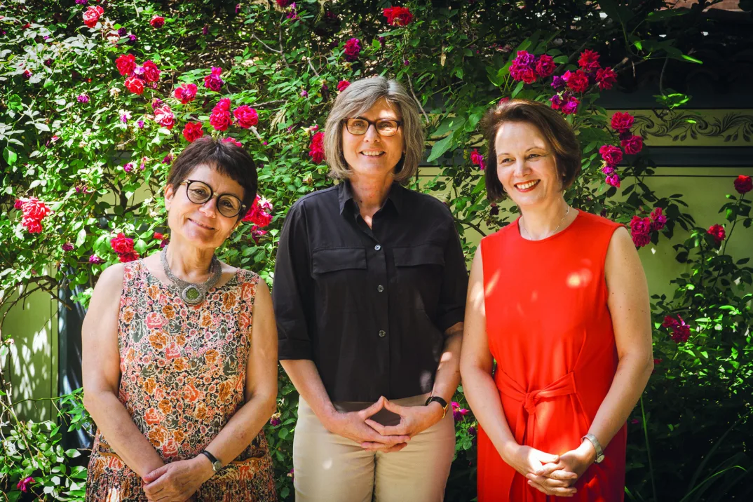 Three women standing smiling in front of a rose bush
