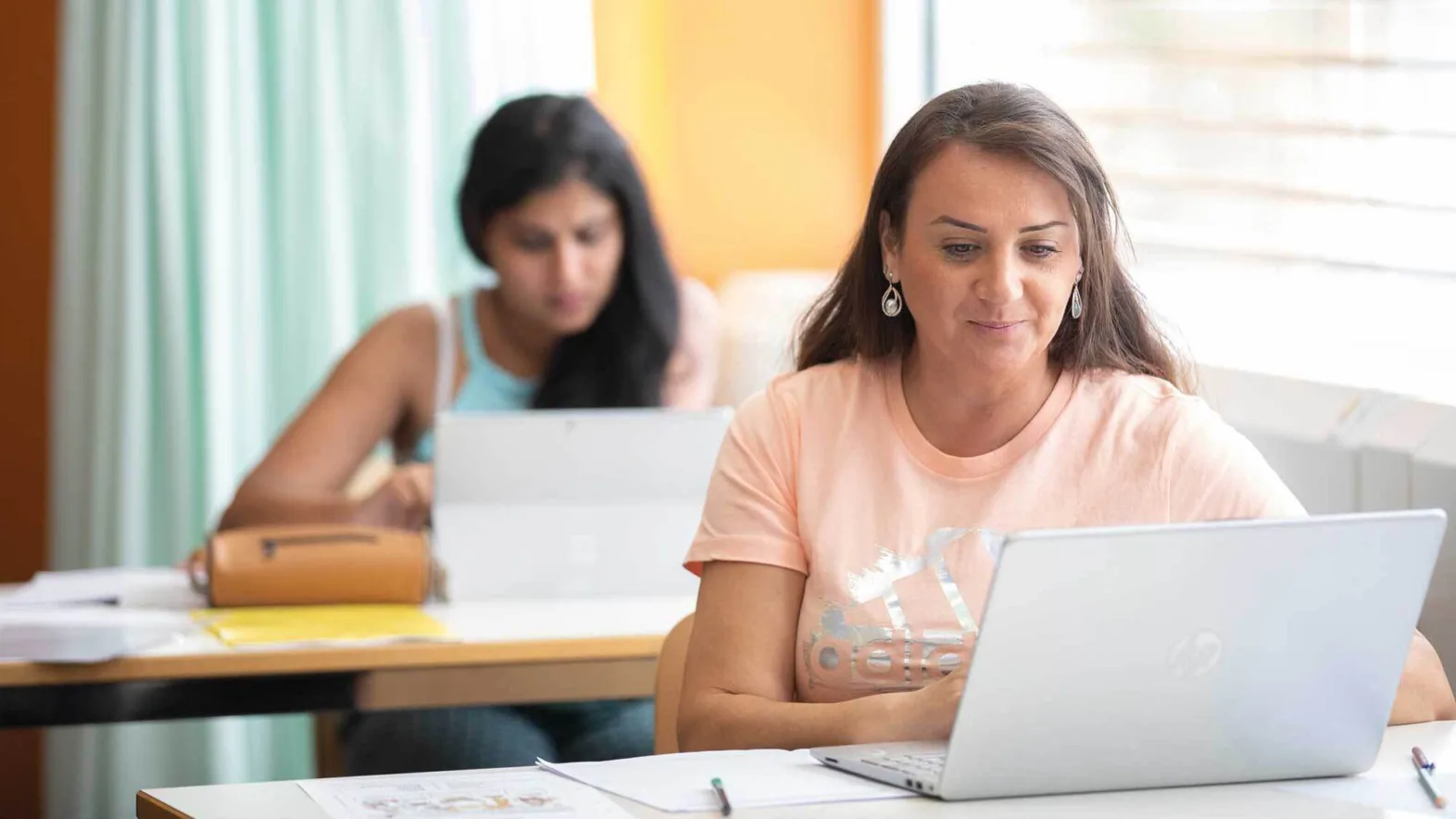 Zwei erwachsene Frauen in der Schule mit einem Laptop vor sich auf dem Schreibtisch.