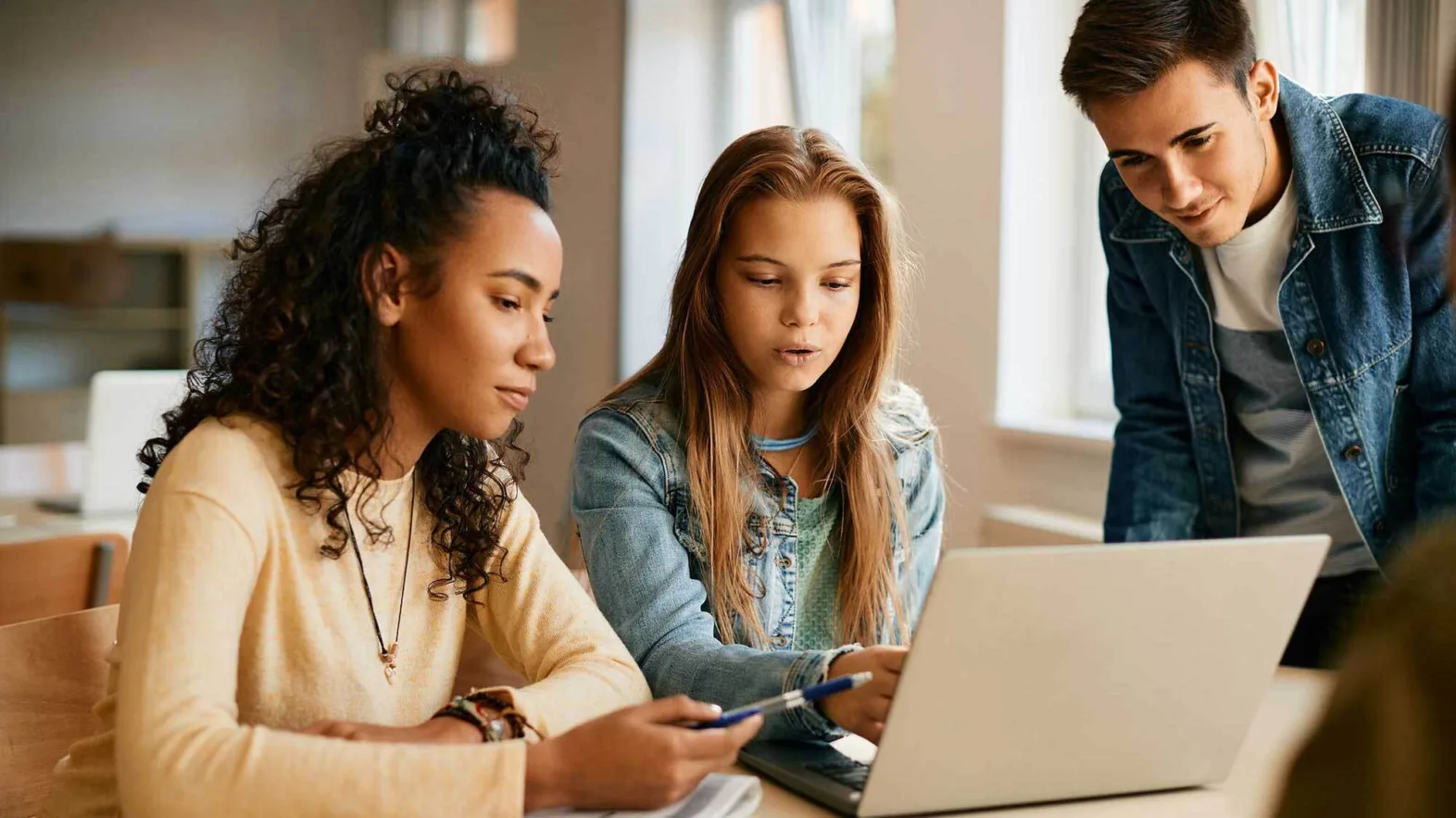 Drei Jugendliche in der Schule schauen auf den Bildschirm eines Notebooks und diskutieren.