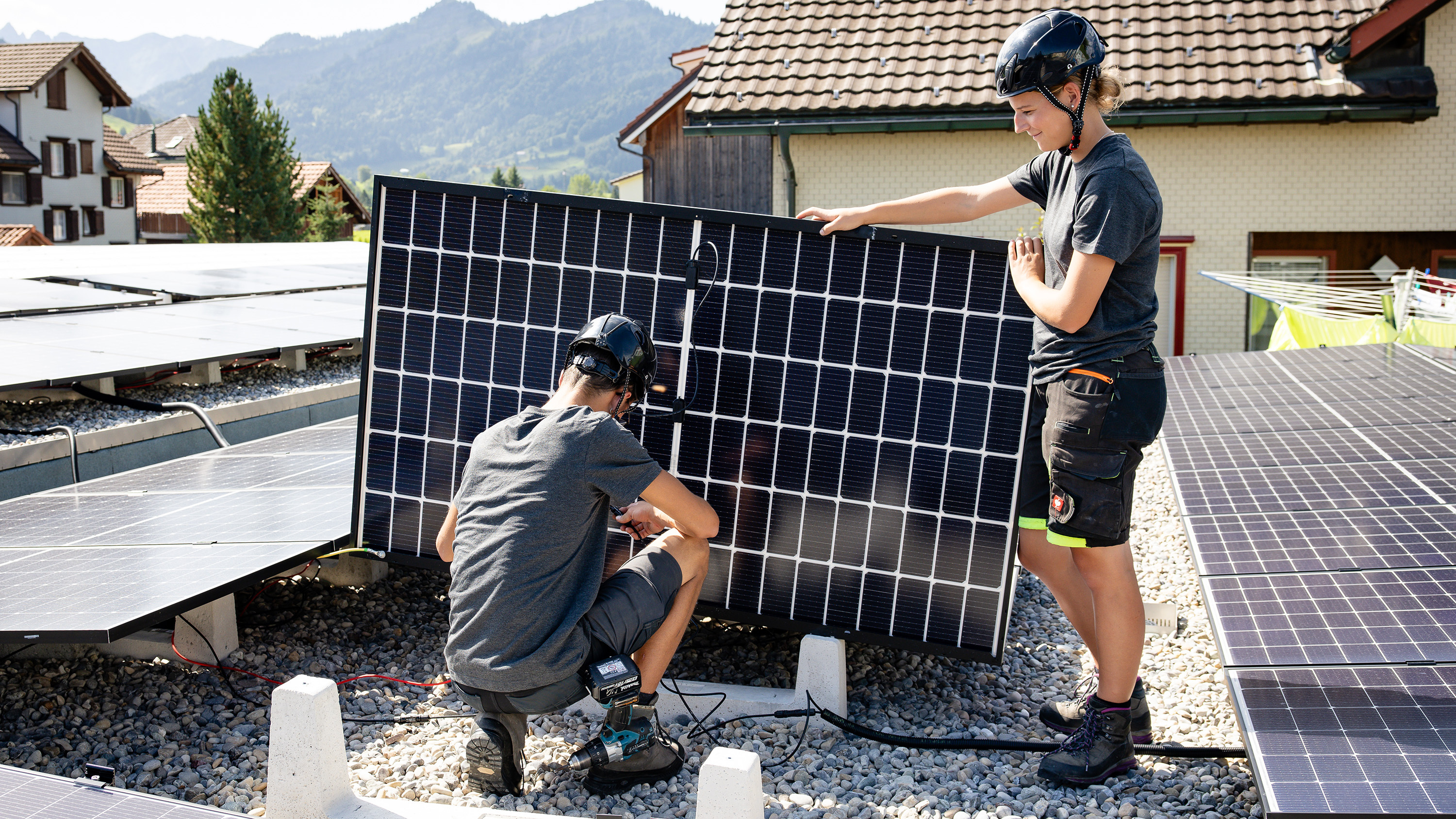 Deux apprentis travaillent sur des panneaux solaires en tenue de travail et avec un casque sur un toit plat.