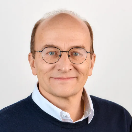 Portrait of Fredy Fritsche, wearing a blue pullover with a white shirt collar and glasses against a light grey background.