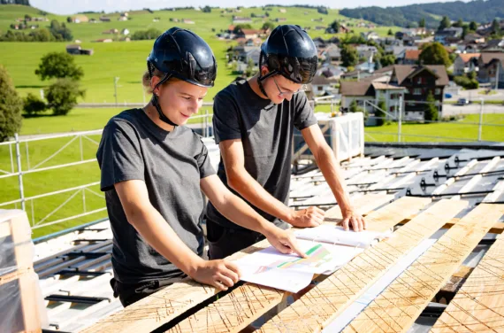 Eine lernende Solarinstallateurin und ein lernender Solarinstallateur sehen sich Pläne auf einer Baustelle auf einem Dach an.