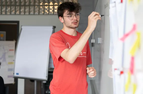 A young man with brown hair, glasses and a red T-shirt is writing something on a whiteboard.