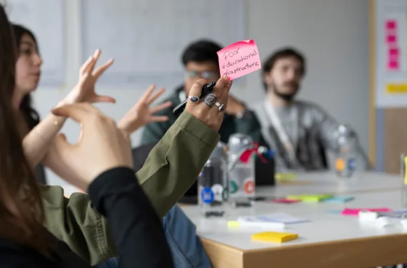 A young woman holds a pink post-it note that reads "poor educational infrastructure". Her team members can be recognised blurred in the background.