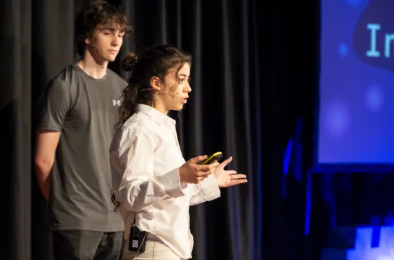 A young woman and a young man are presenting on a stage with a presentation next to them.