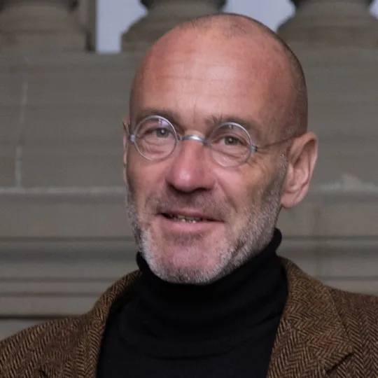 Portrait of a smiling man with a bald head and glasses, wearing a black turtleneck jumper and a brown blazer.