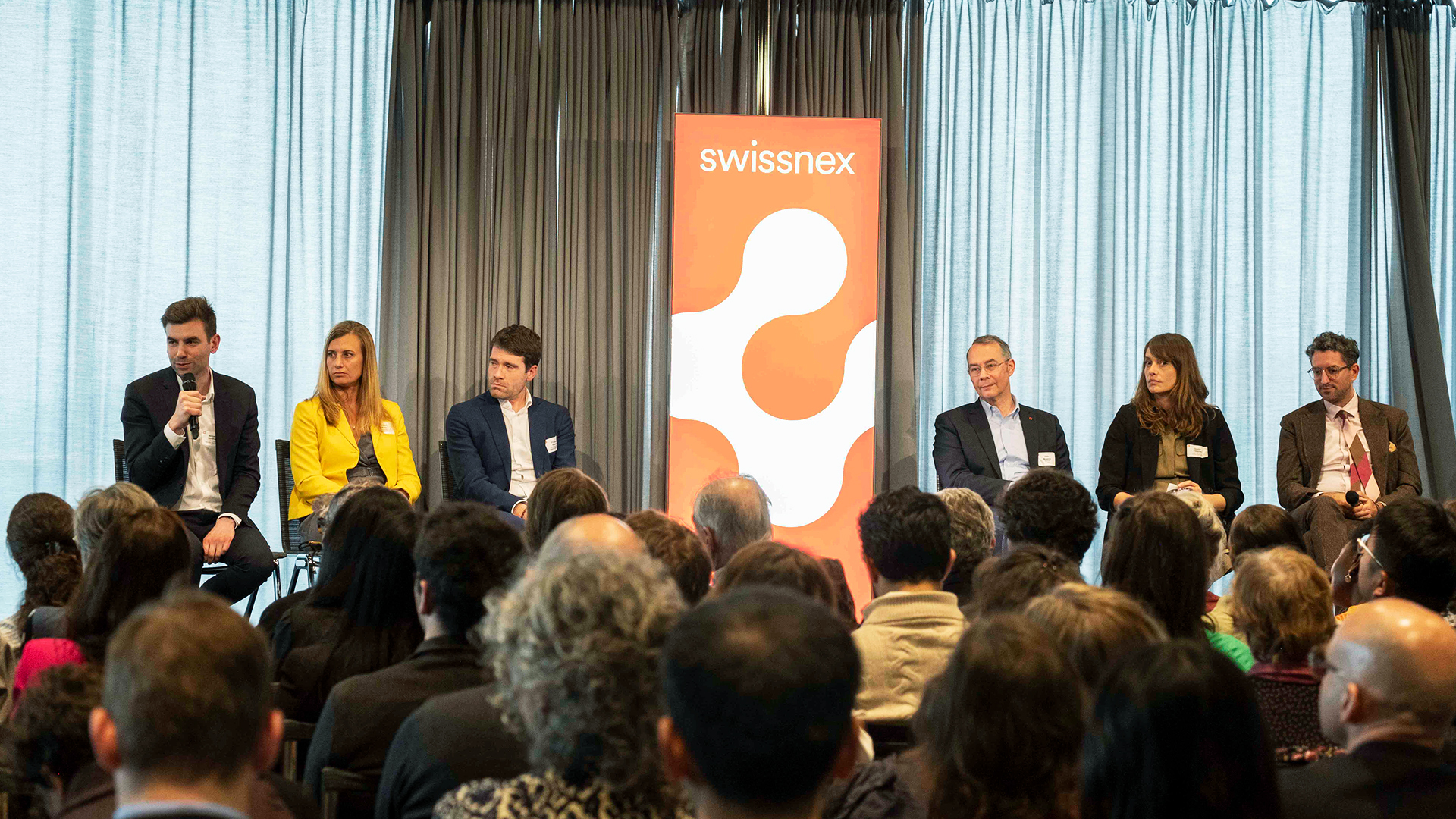 Six Swissnex CEOs are sat on stage around a red banner with the Swissnex logo. The heads of the audience can be seen on the bottom, the background of the stage is gray.