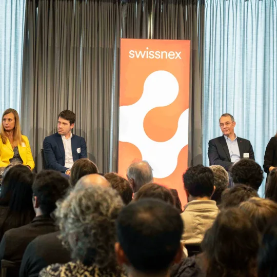Six Swissnex CEOs are sat on stage around a red banner with the Swissnex logo. The heads of the audience can be seen on the bottom, the background of the stage is gray.