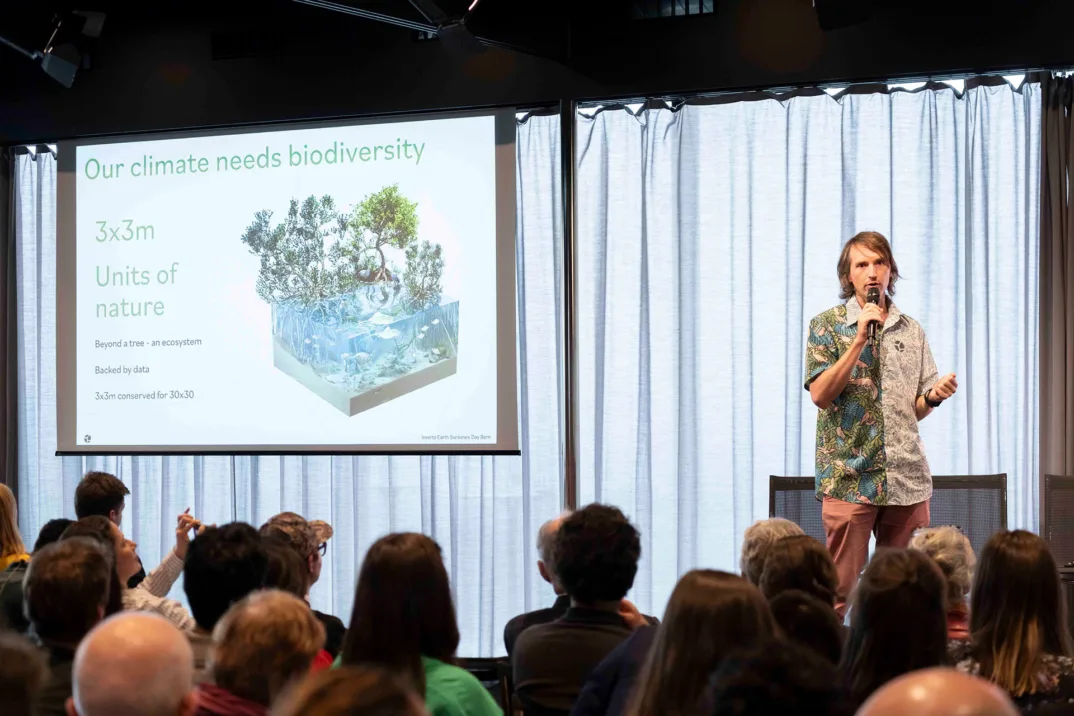 Un homme vêtu d'une chemise à boutons colorée s'adresse sur scène à un public. À côté de lui, un écran contient des informations sur la biodiversité.