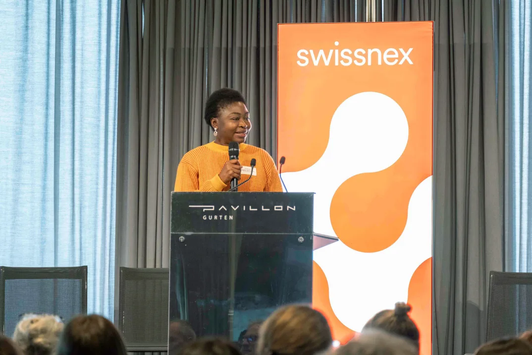 Une femme vêtue d'un pull-over orange s'adresse à un public derrière un podium avec un logo Swissnex en arrière-plan.