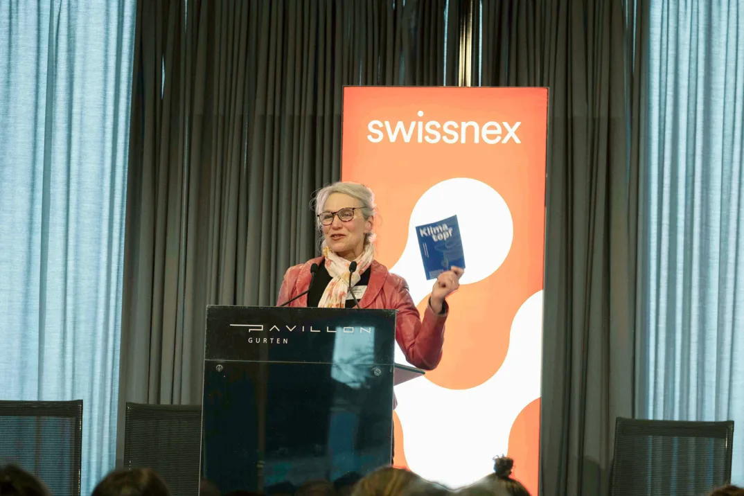 A woman in a red jacket speaks to an audience behind a podium while holding up a leaflet saying "Klimatopf". In the background of the stage is a Swissnex logo.