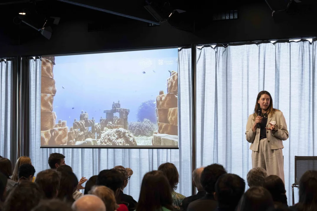 A woman in a beige suit speaks on stage to an audience. Next to her is a screen showing a coral reef.