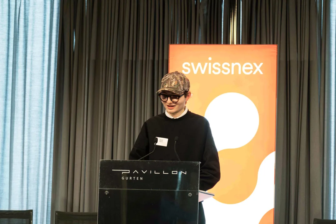 Un homme avec une casquette de baseball et des lunettes s'adresse à un public derrière un podium sur une scène avec un logo Swissnex en arrière-plan.