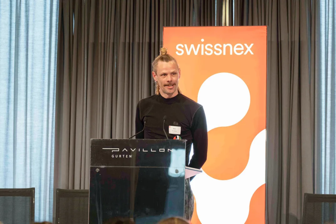 A person speaks behind a podium on a stage to an audience with a Swissnex logo in the background.