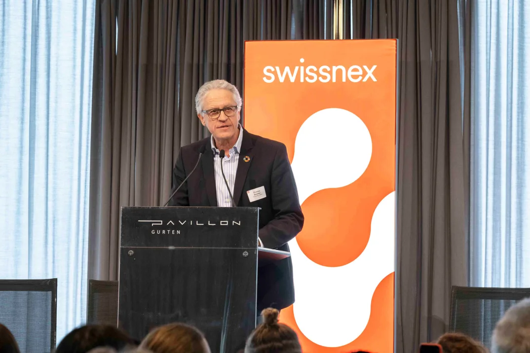 Ein Mann mit Sakko und Brille spricht zu einem Publikum auf einem Podium mit einem Swissnex-Logo im Hintergrund.