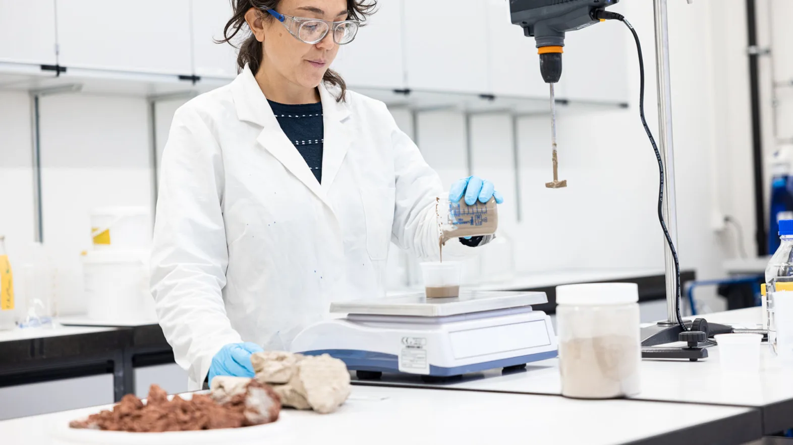A woman wearing a white lab coat, goggles and gloves works in a lab.