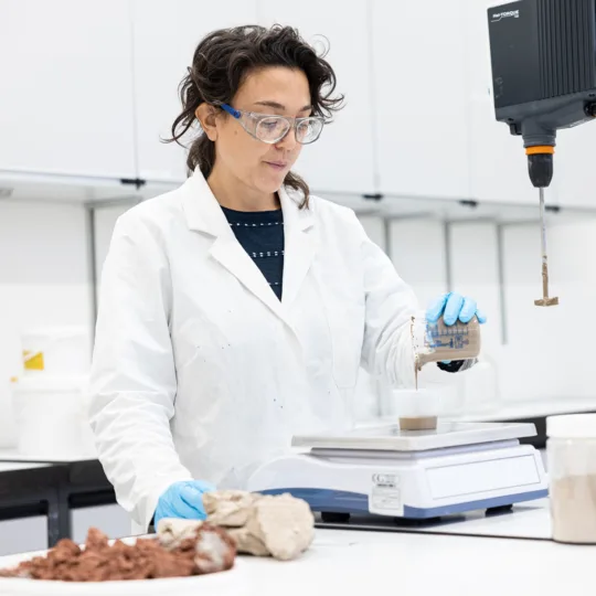 A woman wearing a white lab coat, goggles and gloves works in a lab.