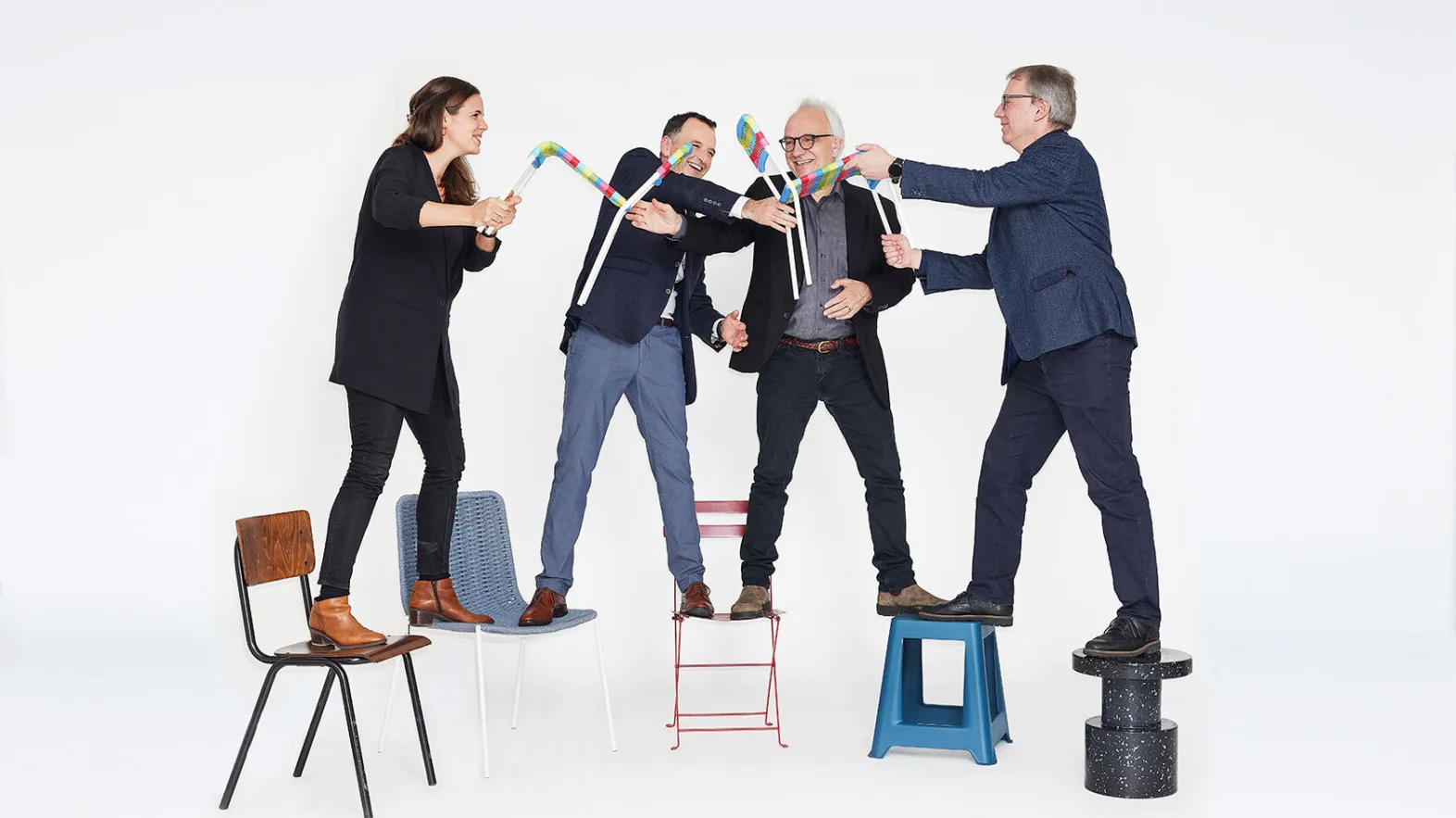 Four people stand with one leg on each on chairs or stools, arranged in a semi-circle and hold up two small, colourful chairs together.