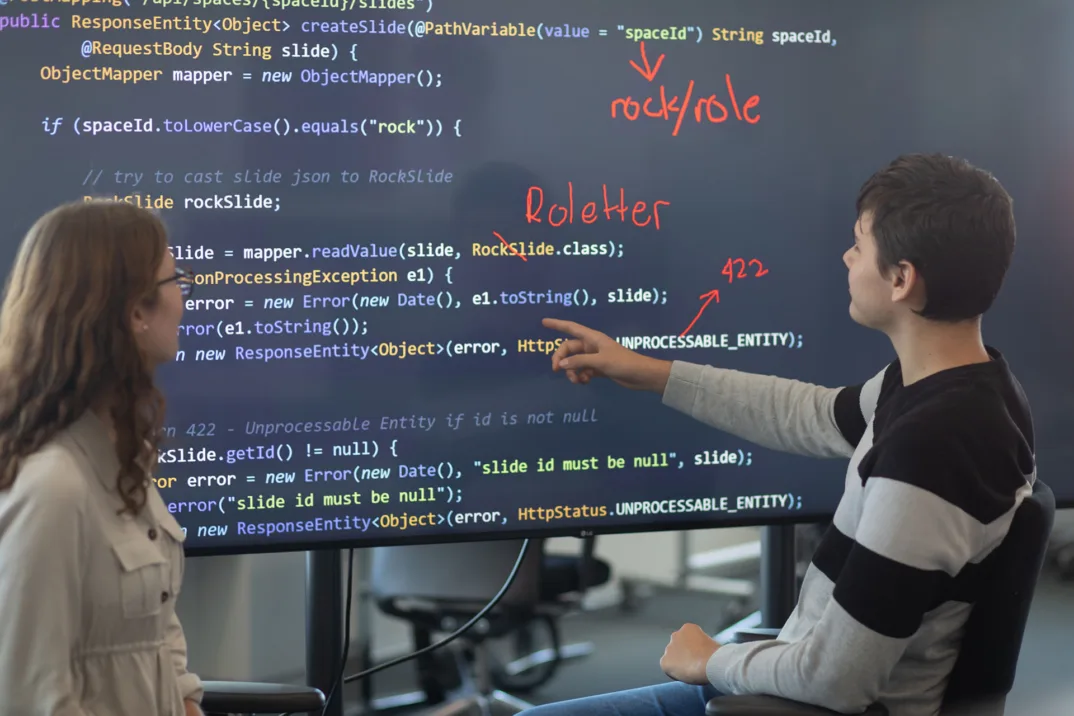 A trainee indicates something to another trainee something on a large screen with code.