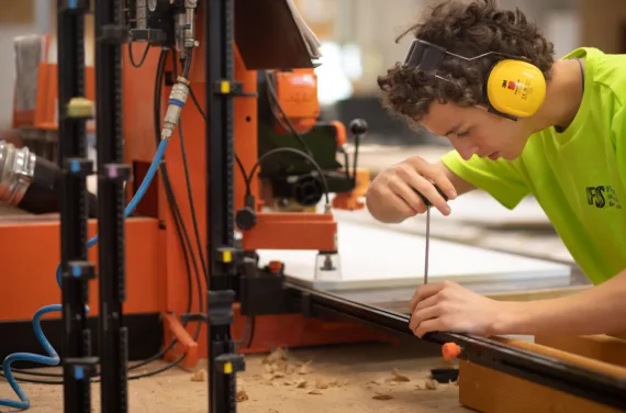 Un apprenti portant un T-shirt vert et des protections auditives jaunes au travail dans une menuiserie.