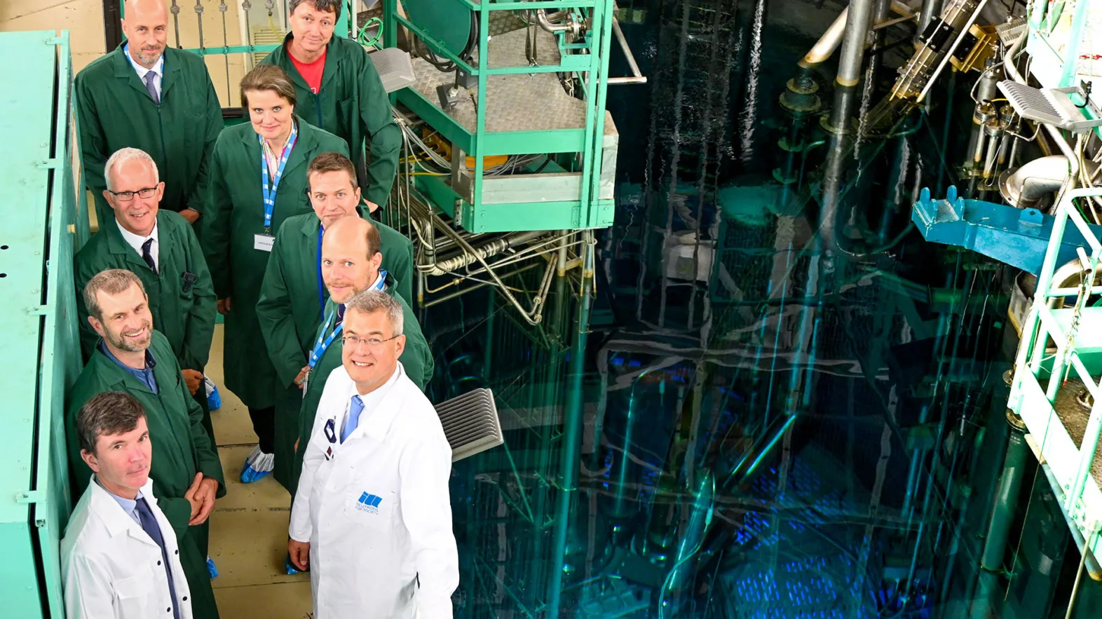 The representatives from SERI and ILL stand in front of a pool at the ILL wearing either green or white coats and looking up towards the camera.