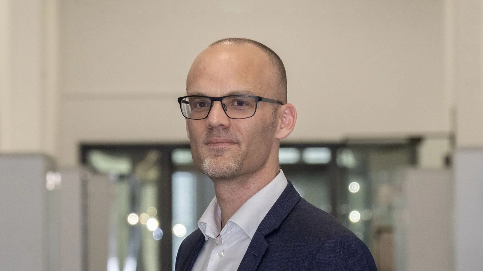 Portrait of a man in a suit wearing glasses in a hallway.