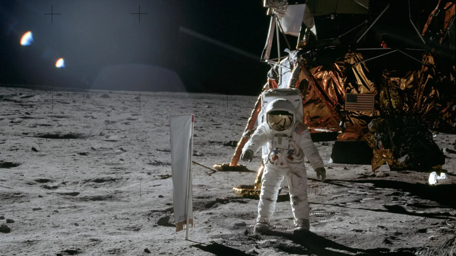 Vor einem Astronauten im Raumanzug auf der grauen Mondoberfläche befindet sich ein weiß-silbernes Tuch an einer Stange. Im Hintergrund ist die dunkle kupferfarbene Mondfähre vor schwarzem Hintergrund.