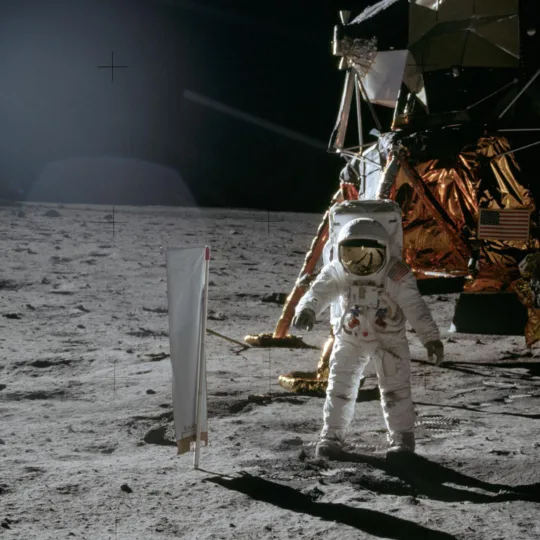 Un drap blanc sur un poteau se trouve devant un astronaute en combinaison spatiale sur la surface de la lune. À l'arrière-plan se trouve le module de commande ombre et cuivré, le fond est noir.