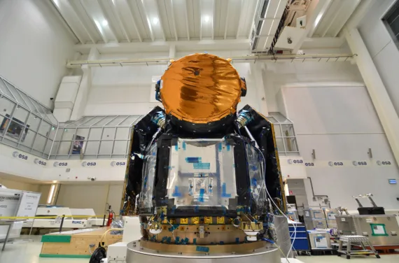 The Cheops telescope in a tall white hall. It is a black and silver cube on the bottom and a copper circle on top.