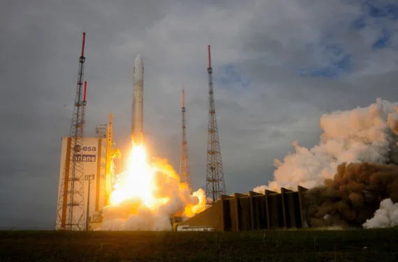 A rocket lifts off into the cloudy and dark sky, a trail of fire below it.