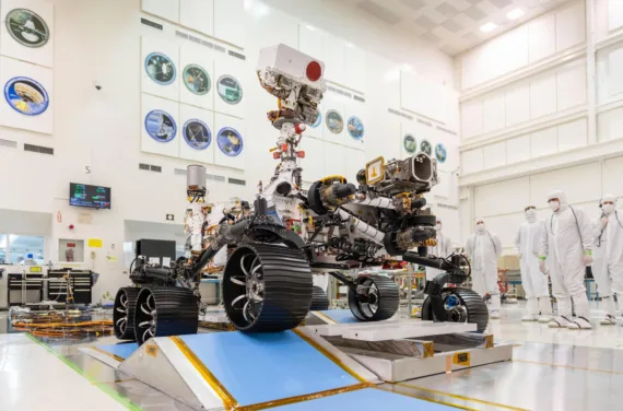 A mars rover drives over an obstacle in a tall white hall. It is observed by people in white coveralls wearing masks.