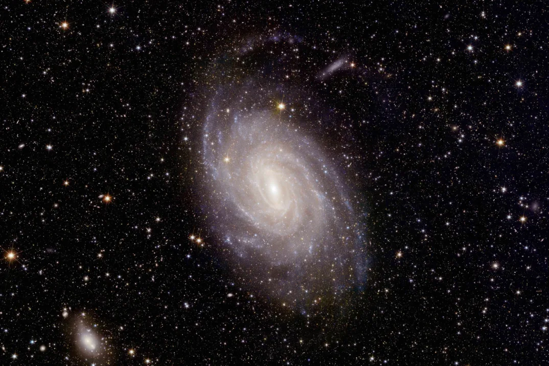 A photo of a galaxy against a dark background. The swirl is white in the center with purple on the outside and it is surrounded my many stars.