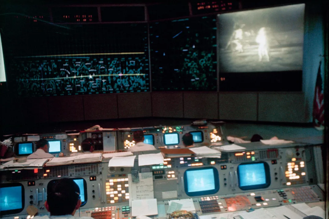 Une vieille photo d'une salle de contrôle de mission sombre, éclairée par de nombreux écrans d'ordinateur à l'avant et une projection de deux astronautes sur la lune à l'arrière.