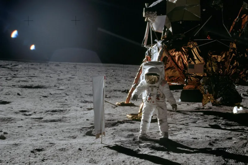 Vor einem Astronauten im Raumanzug auf der grauen Mondoberfläche befindet sich ein weiß-silbernes Tuch an einer Stange. Im Hintergrund ist die dunkle kupferfarbene Mondfähre vor schwarzem Hintergrund.