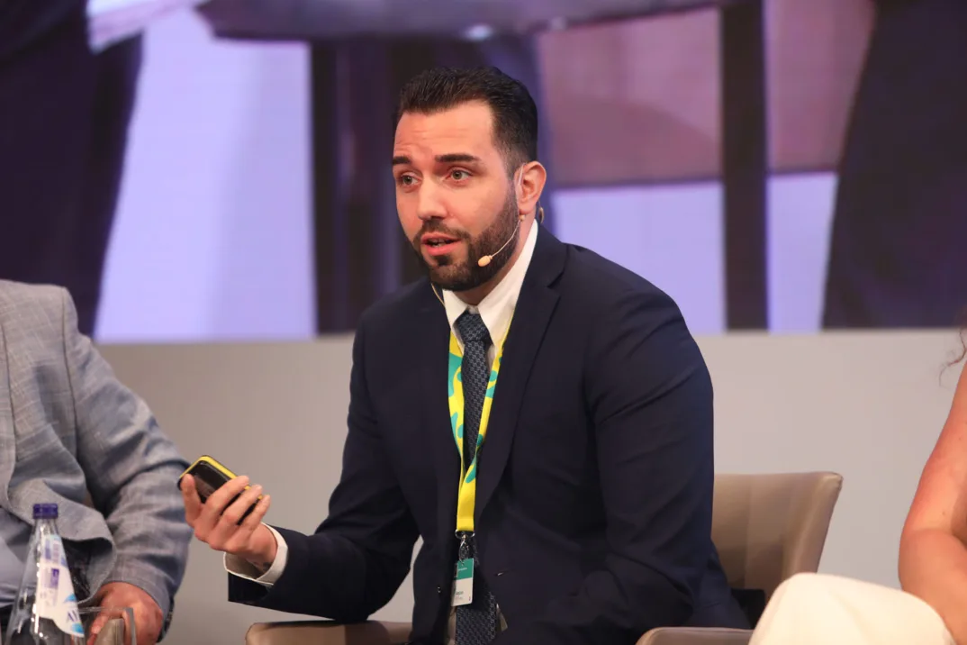 A man in a suit sits on a stage and speaks into a headset microphone.