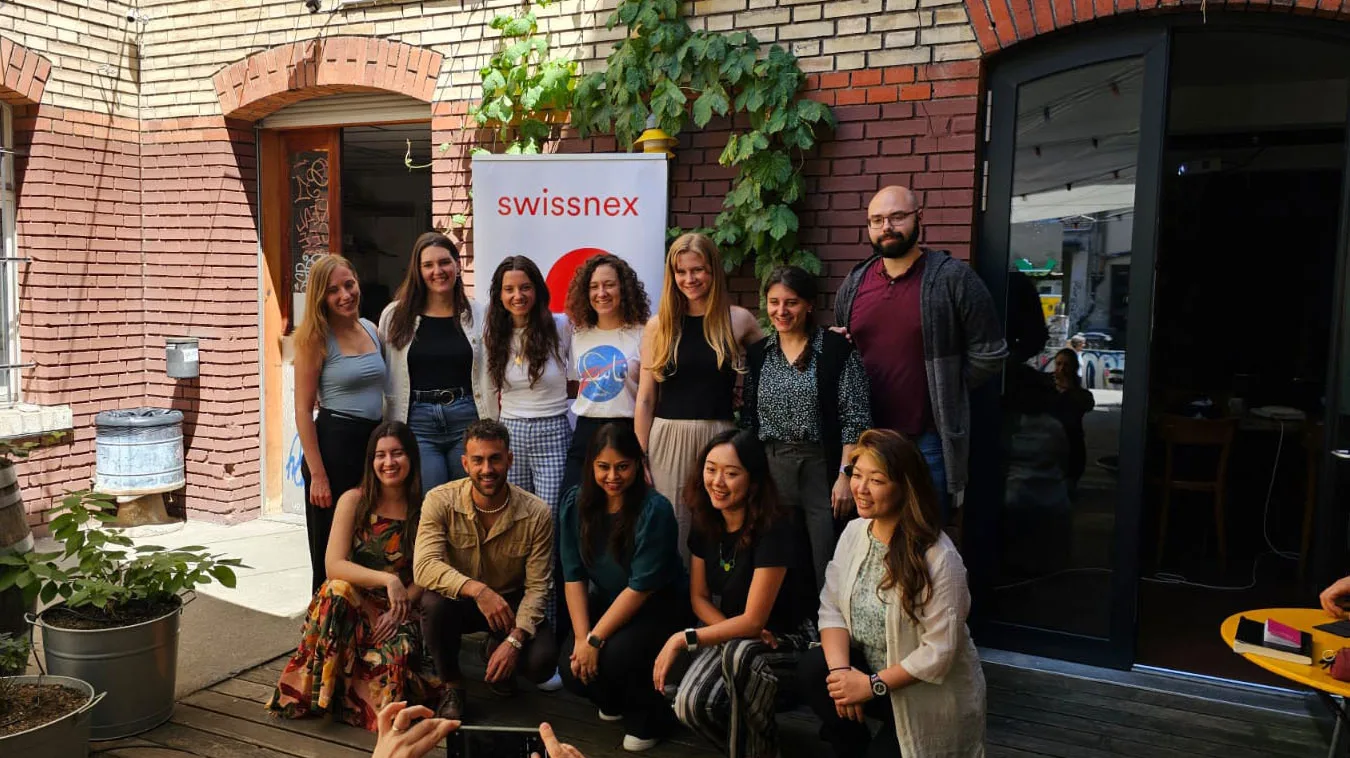 A group of people pose for a picture in a courtyard in front of a brick wall and a Swissnex banner.