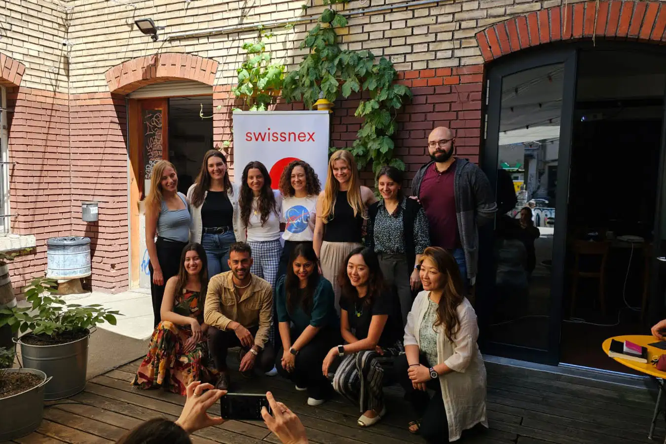 Un groupe de personnes pose pour une photo dans une cour devant un mur de briques et une bannière Swissnex.