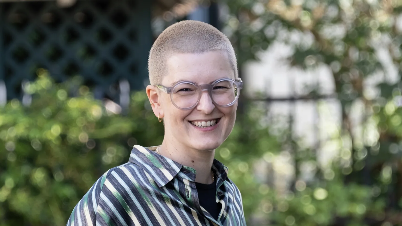 Une femme aux cheveux blonds et courts, portant des lunettes et un chemisier à rayures, sourit à la caméra.