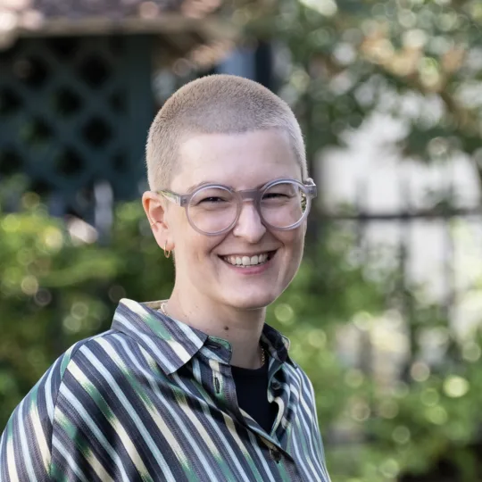 Une femme aux cheveux blonds et courts, portant des lunettes et un chemisier à rayures, sourit à la caméra.