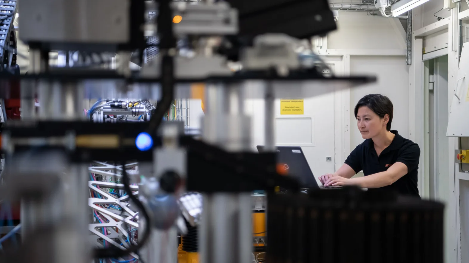 Eine Frau mit kurzen schwarzen Haaren in einem schwarzen T-Shirt arbeitet an einem Laptop umgeben von technischen Geräten.