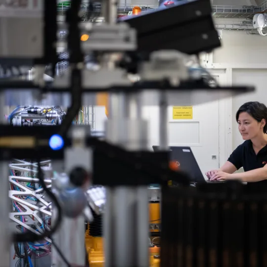 Eine Frau mit kurzen schwarzen Haaren in einem schwarzen T-Shirt arbeitet an einem Laptop umgeben von technischen Geräten.