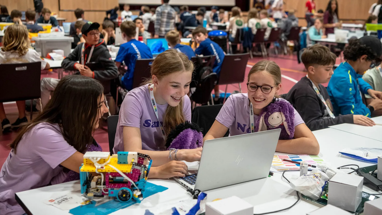 Three young girls in purple T-shirts sit at a laptop in a hall surrounded by other young participants.