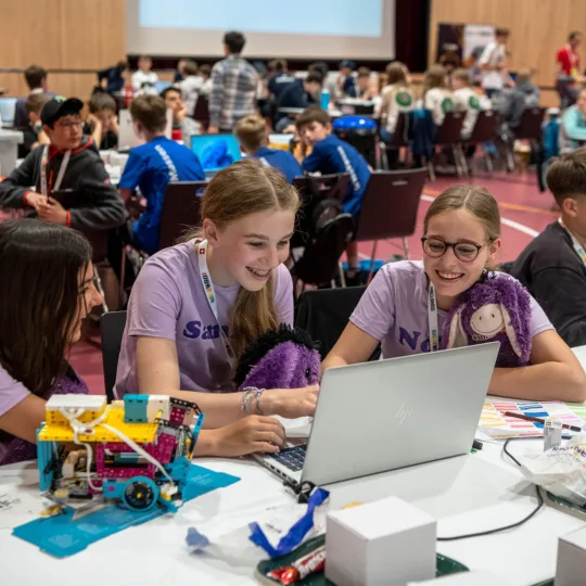 Three young girls in purple T-shirts sit at a laptop in a hall surrounded by other young participants.