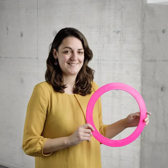 A smiling woman with brown hair in a yellow shirt poses in front of a concrete wall with a large pink ring.