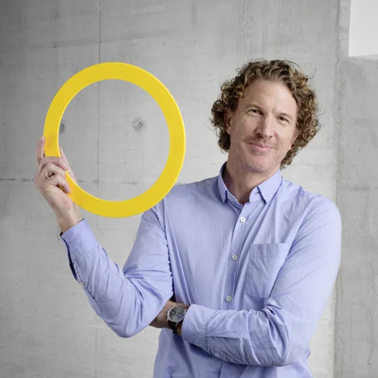 Un homme aux cheveux châtains clairs et à la chemise bleue pose en souriant devant un mur de béton avec un grand anneau jaune.