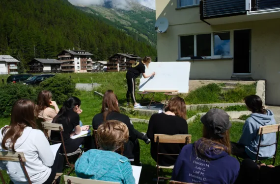 Young people sit in front of a person at a whiteboard in a village in the Alps.