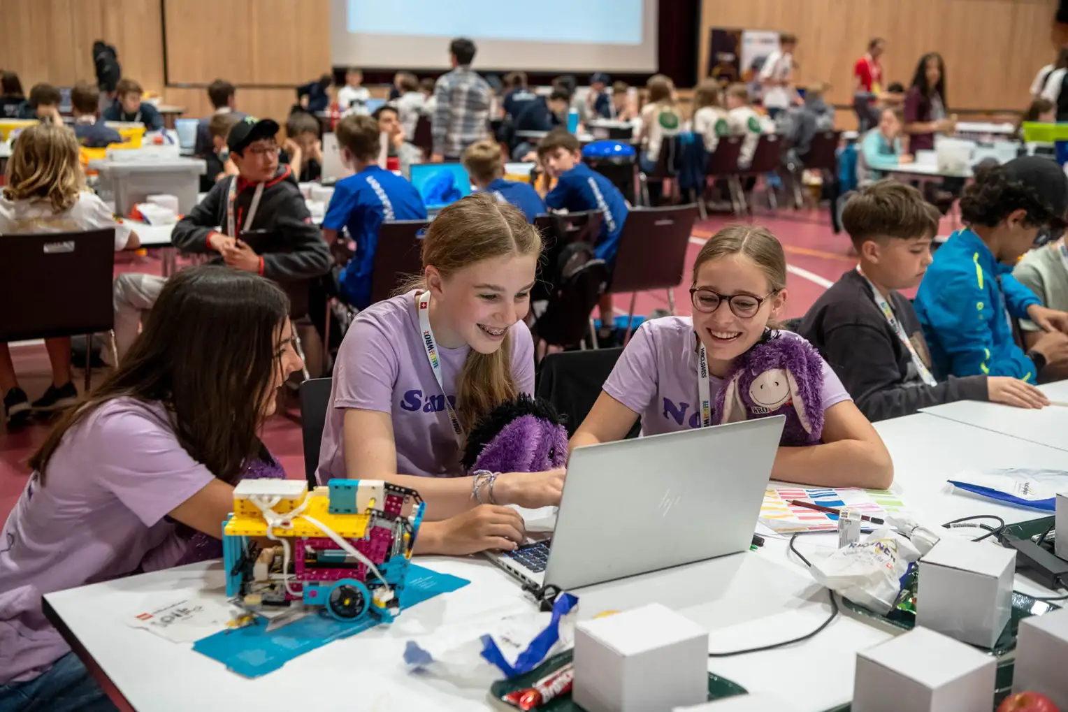 Trois jeunes filles en T-shirt violet sont assises devant un ordinateur portable dans un hall, entourées d'autres jeunes participants.