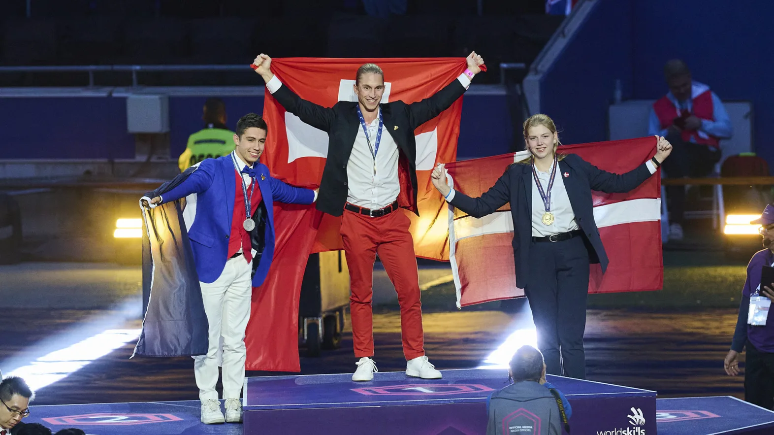 Trois jeunes gens se tiennent sur un podium et tiennent leurs drapeaux nationaux derrière eux: une Autrichienne à la troisième place, un Français à la deuxième place, et un Suisse à la première place.