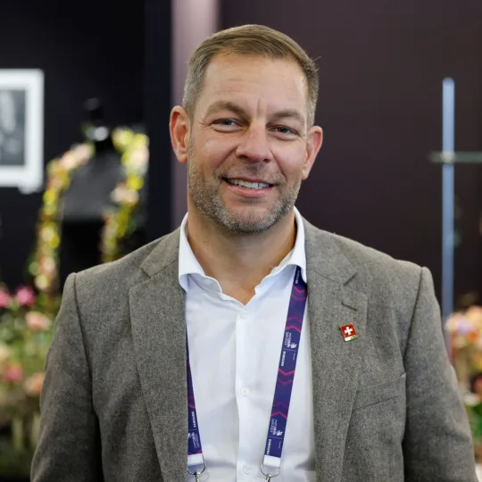 A man in a grey jacket and a white shirt smiles at the camera.