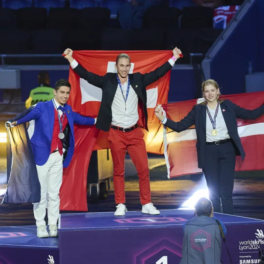 Three young people are standing on a podium, holding the flags of their countries behind them: an Austrian in third place, a Frenchman in second place and a Swiss in first place.